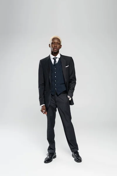 Lunghezza intera di elegante uomo afroamericano in abito formale in piedi con mano in tasca su grigio — Foto stock