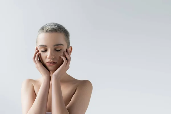 Charmante femme aux épaules nues et aux yeux fermés touchant le visage isolé sur gris — Photo de stock