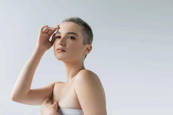 Giovane donna che distoglie lo sguardo mentre tiene la mano vicino al viso isolato sul grigio — Foto stock