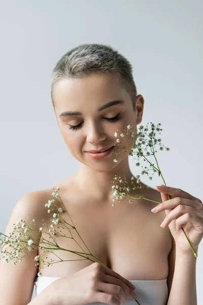 Pretty woman with naked shoulders and closed eyes smiling near branches of gypsophila isolated on grey — Stock Photo