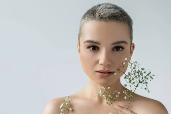 Charming woman with naked shoulders looking at camera near small white flowers isolated on grey — Stock Photo