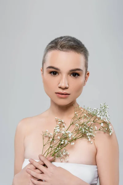 Молода жінка з ідеальною шкірою і білими квітами гіпсофіли дивиться на камеру ізольовано на сірому — стокове фото