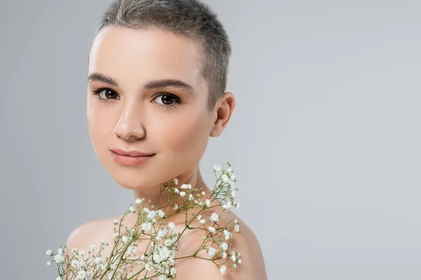 Mujer encantadora con pelo corto y maquillaje natural mirando a la cámara cerca de pequeñas flores blancas aisladas en gris - foto de stock