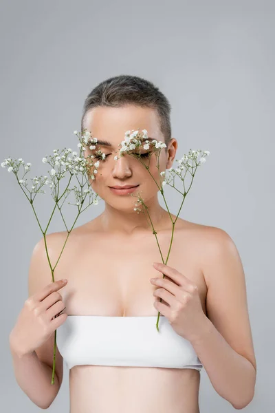 Bonita mujer con los ojos cerrados y hombros desnudos sosteniendo ramas de gypsophila aislado en gris - foto de stock