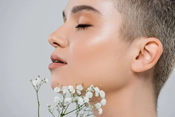 Perfil de mujer encantadora con cara perfecta cerca de pequeñas flores blancas aisladas en gris - foto de stock