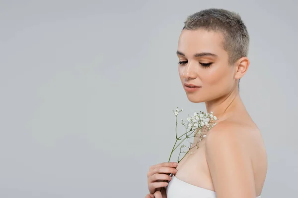 Jolie femme aux cheveux courts et aux épaules nues tenant de minuscules fleurs de gypsophile isolées sur gris — Photo de stock