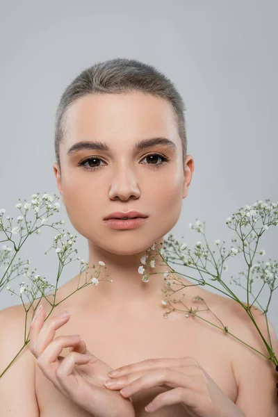 Porträt einer jungen Frau mit natürlichem Make-up, die in die Kamera blickt, in der Nähe weißer winziger Blüten, isoliert auf grau — Stockfoto