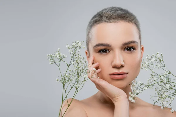 Pretty woman with short hair touching face and looking at camera near white tiny flowers isolated on grey — Stock Photo
