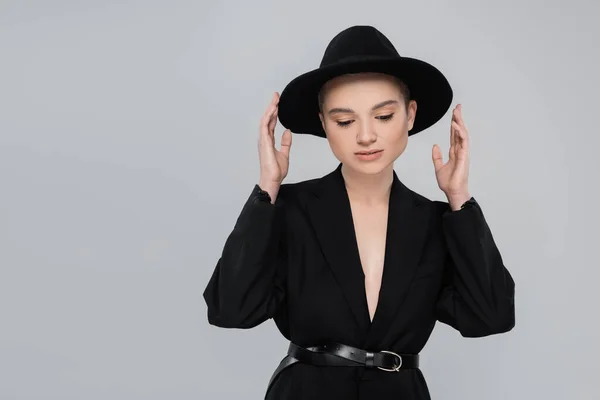 Модна жінка з натуральним макіяжем торкається чорного капелюха ізольовано на сірому — стокове фото