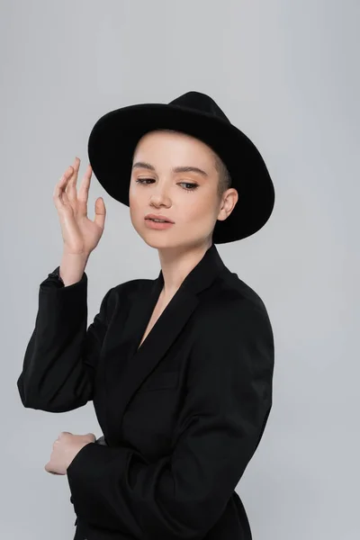 Jeune femme en blazer noir touchant bord chapeau isolé sur gris — Photo de stock