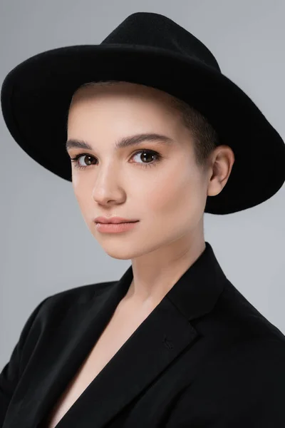 Portrait de jeune femme au maquillage naturel, coiffée d'un chapeau noir, regardant la caméra isolée sur fond gris — Photo de stock