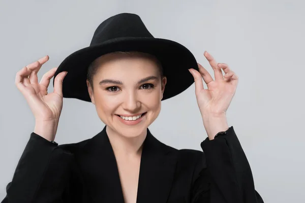 Front view of smiling woman adjusting black brim hat isolated on grey — Stock Photo