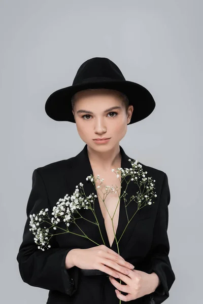 Mulher elegante em chapéu fedora preto olhando para a câmera perto de pequenas flores brancas isoladas em cinza — Fotografia de Stock