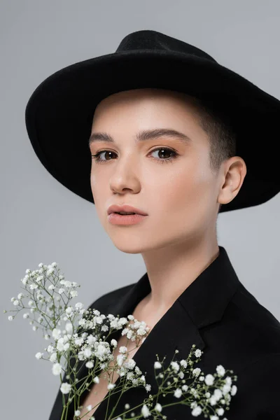 Mujer joven en sombrero fedora negro mirando a la cámara cerca de pequeñas flores blancas aisladas en gris - foto de stock