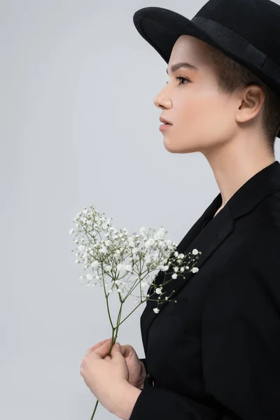 Vista laterale della donna in blazer nero e cappello tesa vicino a fiori di gypsophila bianchi isolati su grigio — Foto stock