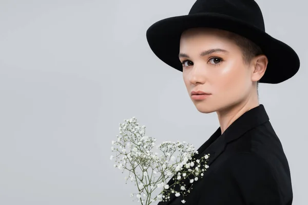 Bella giovane donna in cappello nero fedora guardando la fotocamera vicino a fiori di gypsophila isolati su grigio — Foto stock