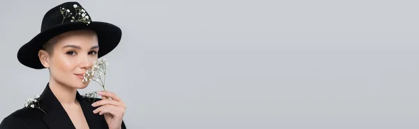 Очаровательная женщина в черной шляпе fedora пахнущие белые крошечные цветы изолированы на серый, баннер — стоковое фото