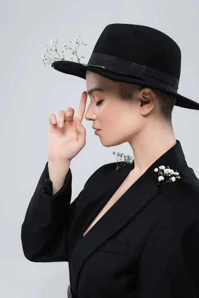 Side view of woman with closed eyes pointing at black brim hat with gypsophila flowers isolated on grey — Stock Photo