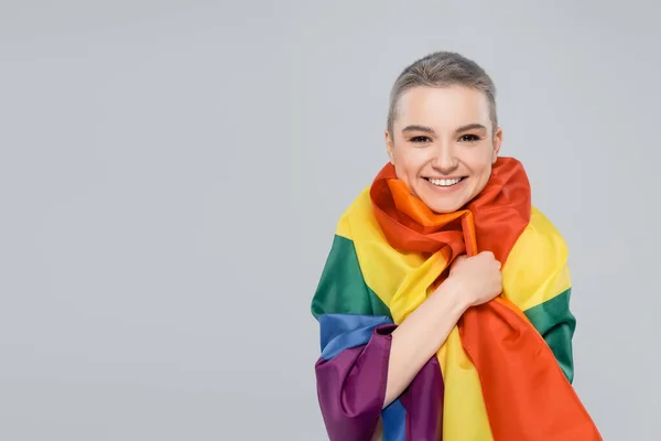 Радісна жінка дивиться на камеру, обгорнувши прапор lgbt ізольовано на сірому — стокове фото