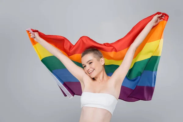 Mulher feliz no topo branco segurando bandeira lgbt em mãos levantadas isolado no cinza — Fotografia de Stock