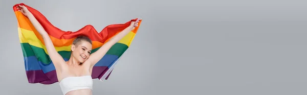 Fröhliche Frau mit lgbt Flagge in erhobenen Händen isoliert auf grau, Banner — Stockfoto