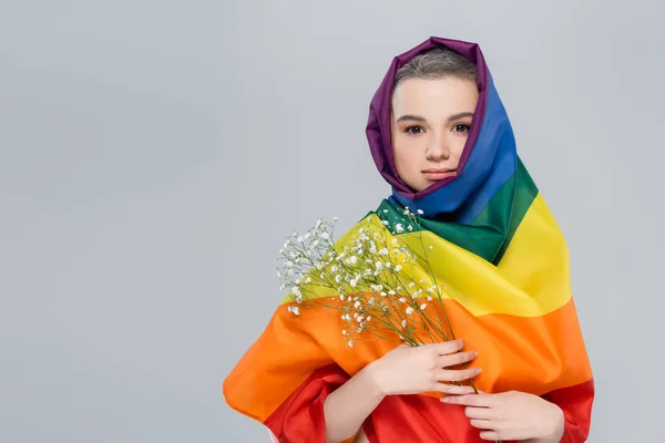 Young woman wrapped in lgbt flag holding gypsophila flowers isolated on grey — Stock Photo