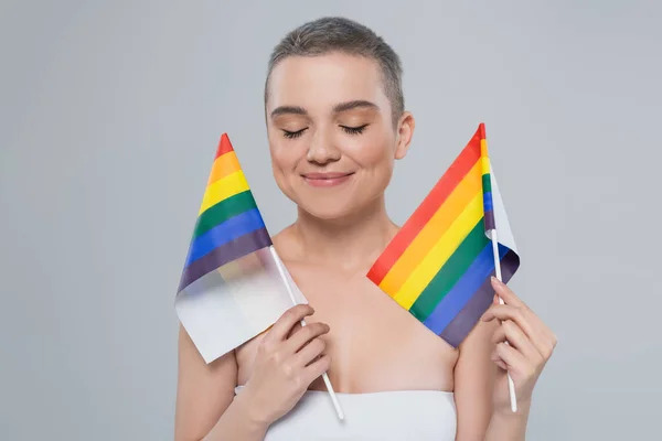 Mulher sorridente com olhos fechados segurando pequenas bandeiras lgbt isolado em cinza — Fotografia de Stock