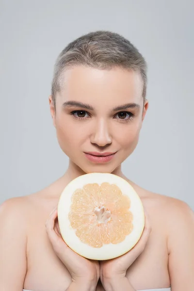 Молода жінка з голими плечима дивиться на камеру біля половини грейпфрута ізольована на сірому — стокове фото
