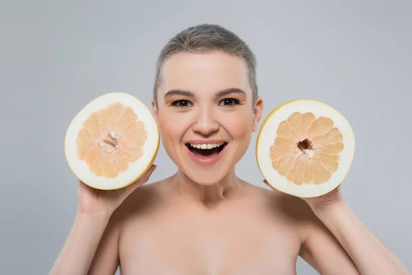 Femme gaie regardant caméra tout en montrant des moitiés de pamplemousse isolé sur gris — Photo de stock