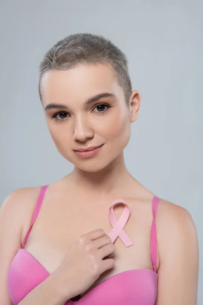 Pretty young woman with breast cancer awareness ribbon looking at camera isolated on grey — Stock Photo