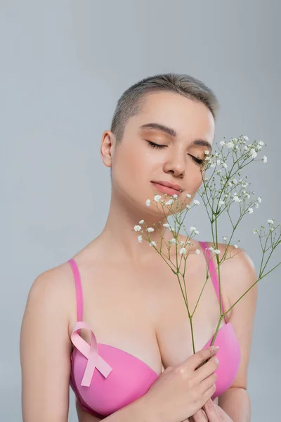 Mulher encantadora com olhos fechados e fita de consciência rosa segurando flores brancas minúsculas isoladas em cinza — Fotografia de Stock