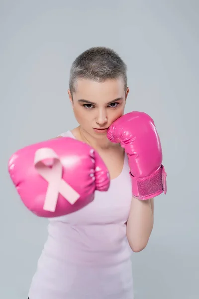 Woman with breast cancer awareness ribbon on pink boxing glove standing in fight position isolated on grey — Stock Photo