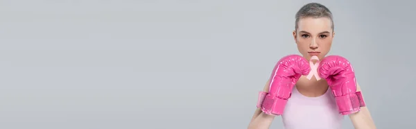 Woman in pink boxing gloves looking at camera and holding breast cancer awareness ribbon isolated on grey, banner — Stock Photo