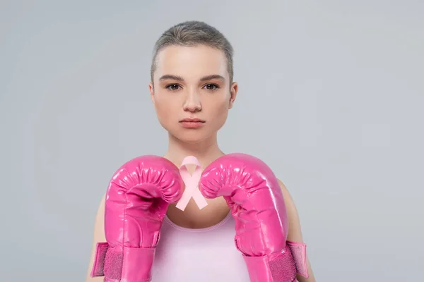 Mujer con el pelo corto, en guantes de boxeo de color rosa, la celebración de cáncer de mama cinta de conciencia aislado en gris - foto de stock