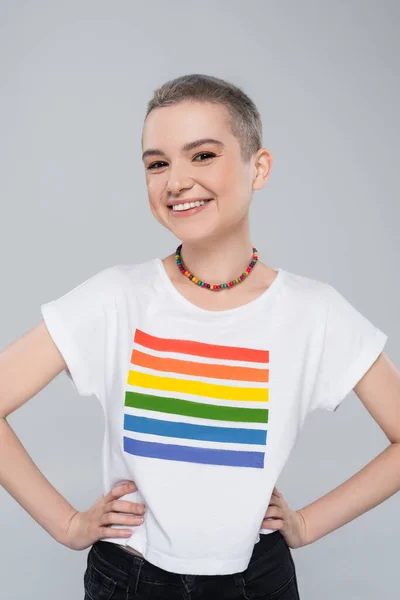 Fröhliche junge Frau in lgbt Farben T-Shirt stehend mit den Händen auf den Hüften isoliert auf grau — Stockfoto