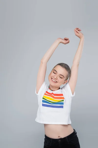 Fröhliche Frau in Regenbogenfarben T-Shirt stehend mit geschlossenen Augen und erhobenen Händen isoliert auf grau — Stockfoto