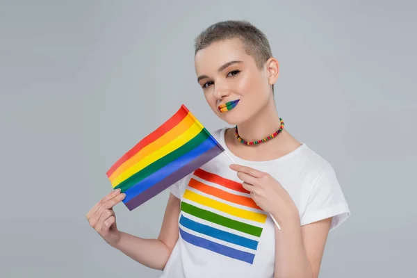 Frau mit regenbogenfarbenen Lippen und kleiner lgbt Flagge, die in die Kamera schaut, isoliert auf grau — Stockfoto