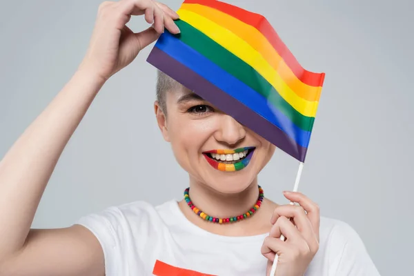 Mujer alegre en colores arco iris collar sosteniendo pequeña bandera lgbt aislado en gris - foto de stock