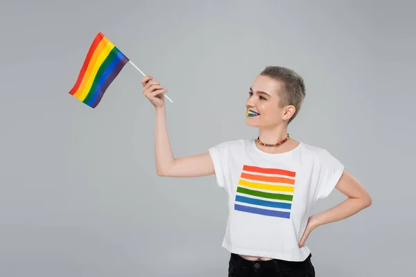 Cheerful woman with small lgbt flag standing with hand on hip isolated on grey — Stock Photo