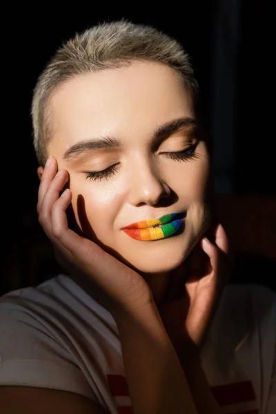 Young woman with rainbow colors lips and hands near face isolated on black — Stock Photo