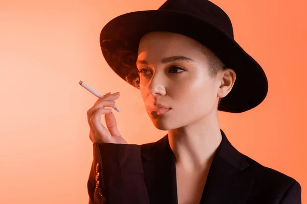 Mujer joven en sombrero fedora negro mirando hacia otro lado mientras fuma sobre fondo de coral - foto de stock