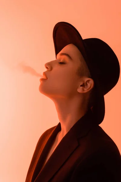 Vista laterale di giovane donna in cappello tesa nera espirando fumo su sfondo corallo — Foto stock