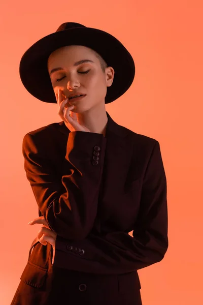 Mujer elegante con los ojos cerrados tocando la cara mientras está de pie aislado en coral - foto de stock