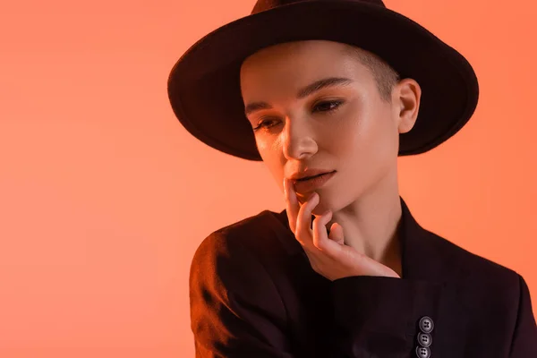 Mujer sensual con maquillaje natural, en sombrero fedora negro, posando con la mano cerca de la cara aislada en coral - foto de stock