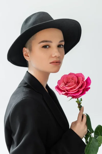 Charming woman with natural makeup, wearing black fedora hat, holding pink rose isolated on grey — Stock Photo