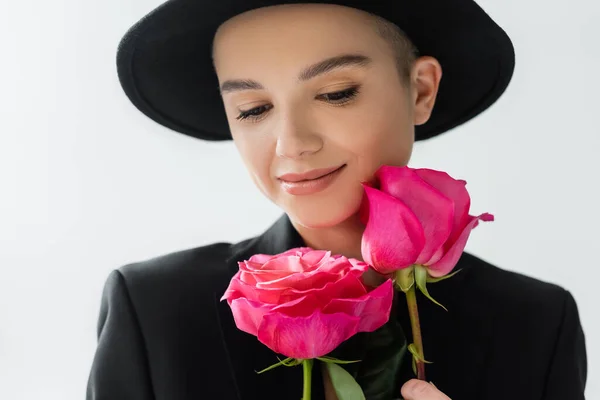 Jovem mulher de chapéu de aba preta e blazer sorrindo perto de rosas rosa isolado em cinza — Fotografia de Stock