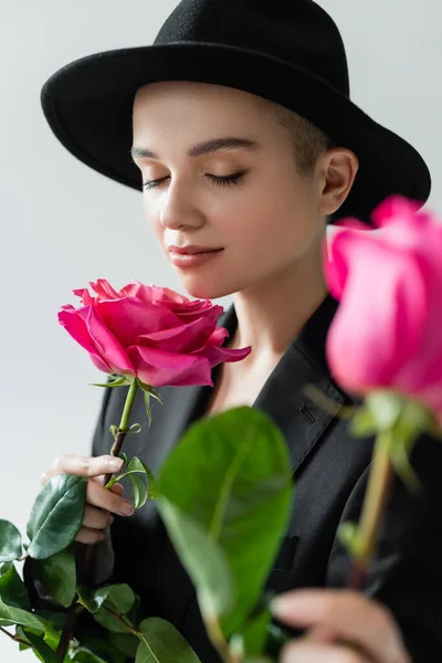 Woman in black brim hat, with closed eyes, smelling aromatic rose on blurred foreground isolated on grey — Stock Photo