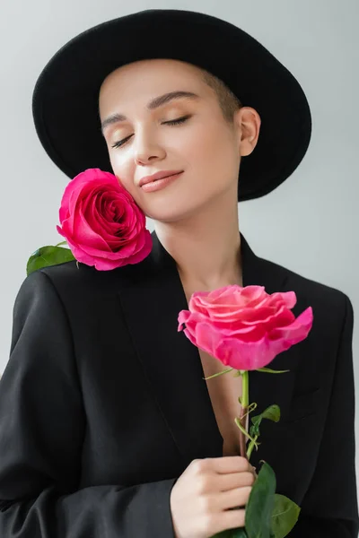 Graziosa donna ad occhi chiusi, in blazer nero e cappello ad ala, in posa con rose rosa isolate su grigio — Foto stock