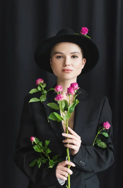 Jeune femme avec un maquillage naturel, portant chapeau bord et blazer, posant avec des roses roses sur noir — Photo de stock