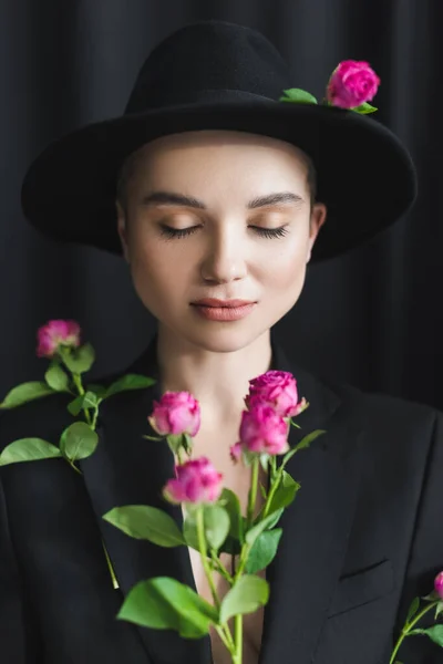 Donna elegante con gli occhi chiusi vicino a rose rosa sfocate su sfondo nero — Foto stock